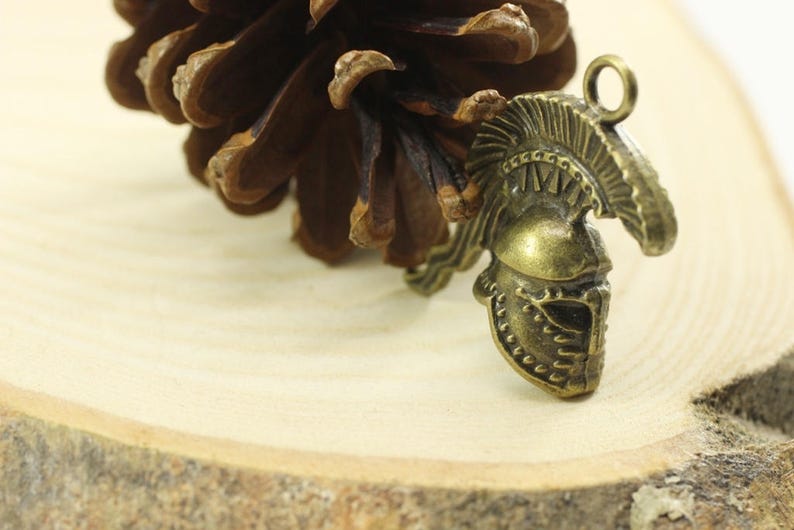 May include: A bronze-colored metal charm in the shape of a Roman helmet with a decorative crest. The charm is on a wooden surface.