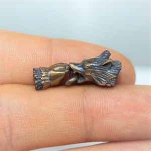Op de afbeelding: Een bronzen metalen bedel met een wolfskop en een hand die een ring vasthoudt. De wolfskop is naar rechts gericht en de hand naar links. De bedel is een gedetailleerd sieraad.