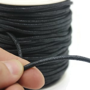 May include: A spool of black waxed cotton cord. The cord is wound tightly around the spool and is being held in a hand.