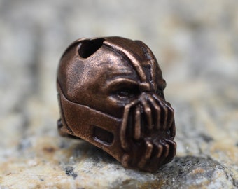 Op de afbeelding: Een koperkleurig metalen beeldje van een personage dat een masker draagt met een gedetailleerd ontwerp. Het masker heeft een scherpe, hoekige vorm en een prominente kaaklijn.