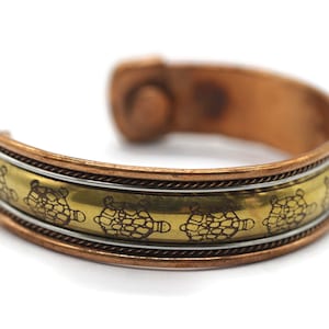 Puede incluir: Un brazalete de cobre y latón con un diseño de tortugas. El brazalete tiene un cierre magnético.