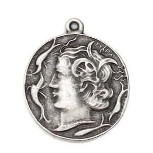 May include: Silver pendant featuring a profile of a woman's face with flowing hair and intricate details. The pendant is circular and has the word "EYRPA" inscribed on the edge.