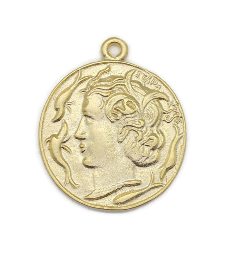 May include: Gold-toned metal pendant featuring a profile of a woman's face with intricate details. The pendant is circular and has a loop at the top for attaching to a chain.