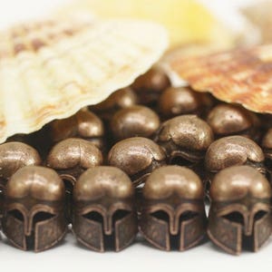 May include: A group of small, bronze-colored metal beads with a detailed design resembling a Spartan helmet. The beads are arranged in a semi-circle on a white surface.