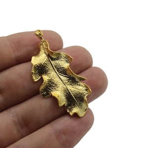 Puede incluir: Un colgante de hoja de roble de metal dorado. La hoja es detallada y realista.