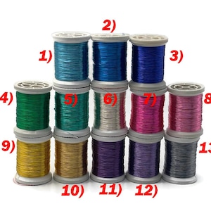 Può includere: Una collezione di tredici rocchetti di filo metallico in vari colori, tra cui verde acqua, blu, viola, verde, argento, rosa, oro e nero. I rocchetti sono disposti in una formazione impilata, mostrando la gamma di colori disponibili.