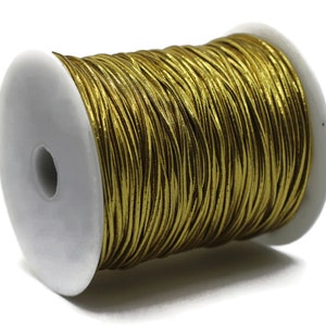 May include: A spool of gold-colored cord wound around a white plastic core. The cord has a metallic sheen and appears to be made of thin, flexible strands. The spool is set against a plain white background.