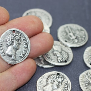 May include: Silver-colored coin pendants with a hole for jewelry making. The coins feature a profile of a man with a laurel wreath and inscriptions. Several coins are visible, some with a hole for stringing.