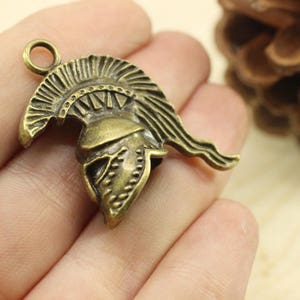 May include: A bronze-colored metal charm in the shape of a Greek warrior helmet. The helmet has a detailed design with a plume on top and a face mask.