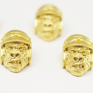 May include: Five gold-colored metal gorilla head charms. Each charm features a detailed gorilla face with a furrowed brow and a slightly open mouth. The charms are all facing forward and are arranged in a row.