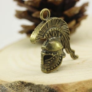 May include: A bronze-colored metal charm in the shape of a Roman helmet with a plume. The helmet has intricate details and a loop at the top for attaching to a necklace or bracelet.