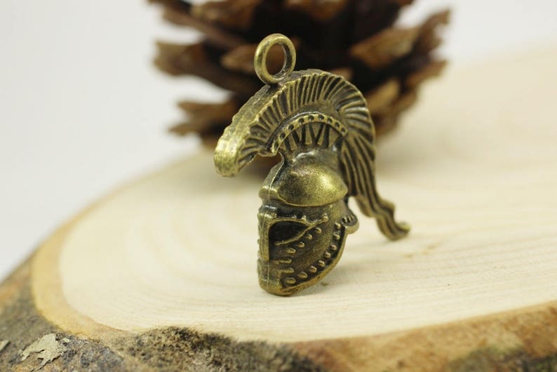 May include: A bronze-colored metal charm in the shape of a Roman helmet with a plume. The helmet has intricate details and a loop at the top for attaching to a necklace or bracelet.
