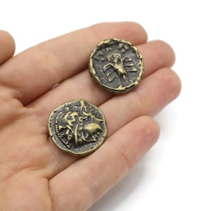 Puede incluir: Dos monedas de color dorado de aspecto antiguo con diseños en relieve. Las monedas muestran un pájaro y un león.