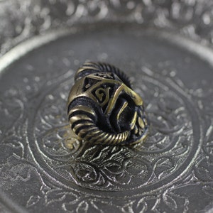 May include: A gold-colored metal bead with a detailed design of a horned helmet. The bead is sitting on a silver tray with an ornate design.