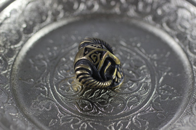 May include: A gold-colored metal bead with a detailed design of a horned helmet. The bead is sitting on a silver tray with an ornate design.