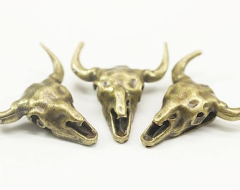 May include: Three antique gold-toned bull skull charms. The skulls are detailed with horns and have an open mouth design.