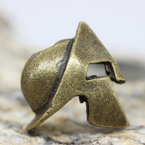 Op de afbeelding: Een kleine, bronzen Spartanen helm bedel. De helm is gedetailleerd met een getextureerd oppervlak en een reli&euml;f ontwerp.