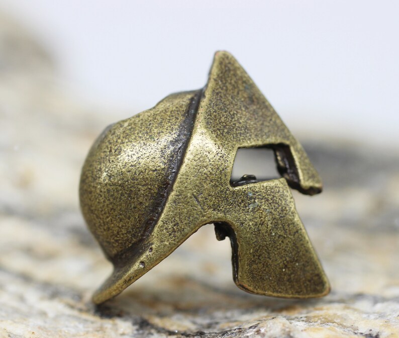 Op de afbeelding: Een kleine, bronzen Spartanen helm bedel. De helm is gedetailleerd met een getextureerd oppervlak en een reli&euml;f ontwerp.