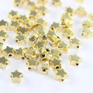 May include: A pile of gold star-shaped beads with a hole in the center of each star. The beads are scattered on a white surface.