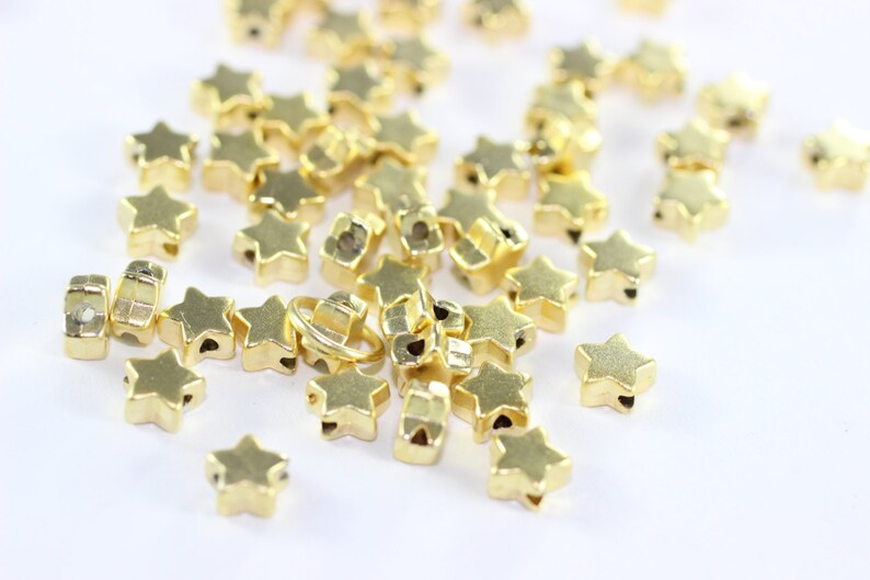 May include: A pile of gold star-shaped beads with a hole in the center of each star. The beads are scattered on a white surface.