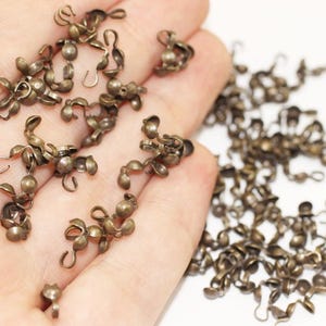 Peut inclure: Une grande quantité de petites attaches métalliques de couleur or antique avec une boucle et une boule à l'extrémité. Les attaches sont éparpillées sur une surface blanche et dans une main.