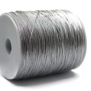 Può includere: Un rocchetto di cordoncino metallico argentato. Il cordoncino è avvolto strettamente attorno al rocchetto, che è fatto di plastica bianca.