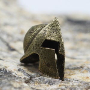 Op de afbeelding: Een klein, bronskleurig Spartaans helm met een getextureerd oppervlak. De helm heeft een puntige kam en een vizier met een kleine opening voor de ogen.