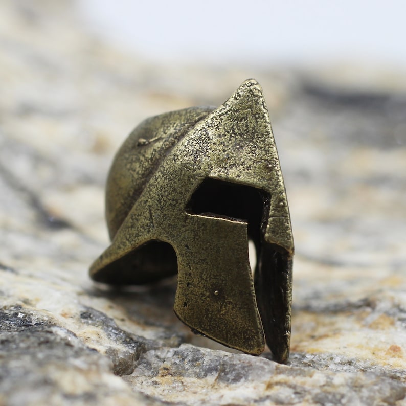 Op de afbeelding: Een klein, bronskleurig Spartaans helm met een getextureerd oppervlak. De helm heeft een puntige kam en een vizier met een kleine opening voor de ogen.