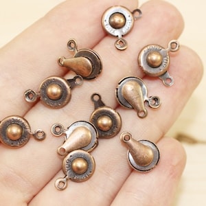 Peut inclure: Plusieurs fermoirs antiques de couleur bronze sont présentés. Chacun possède une base circulaire, un bouton central et un levier. Ces fermoirs, avec de petites boucles d'attache, sont probablement destinés à la bijouterie ou aux travaux manuels.