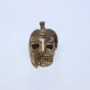 May include: A gold-colored metal skull pendant with a helmet design. The skull has a detailed face with a helmet covering one side of the head.