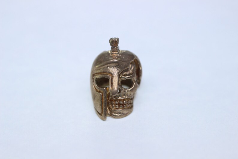 May include: A gold-colored metal skull pendant with a helmet design. The skull has a detailed face with a helmet covering one side of the head.