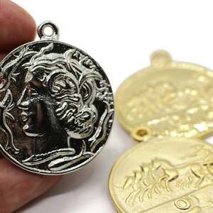 May include: Silver and gold tone metal pendant charms featuring a woman's profile on one side and a chariot with horses on the other.
