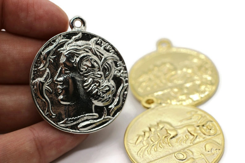 May include: Silver and gold tone metal pendant charms featuring a woman's profile on one side and a chariot with horses on the other.
