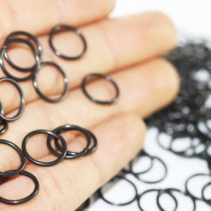 May include: A close-up of a hand holding a large quantity of small, black metal jump rings. The rings are all the same size and are arranged in a random pattern.