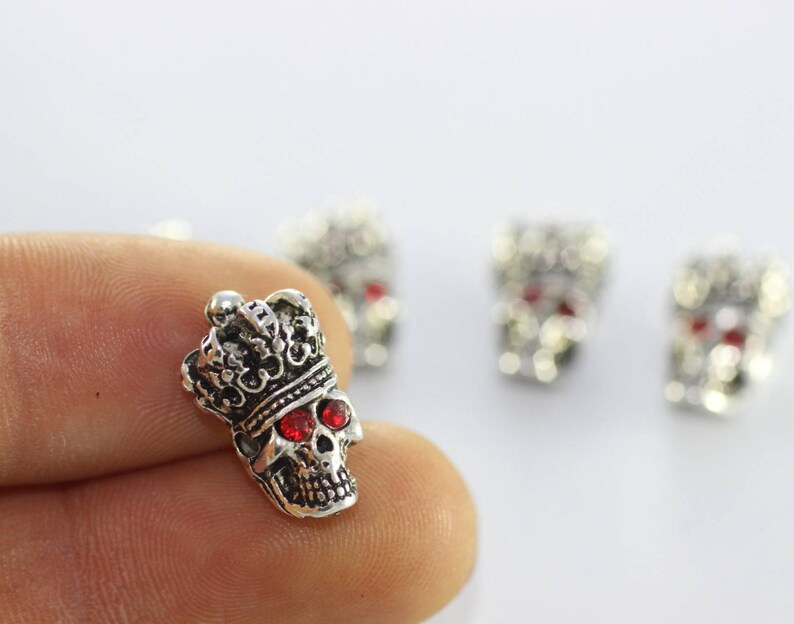 May include: Silver skull charm featuring a crown and red rhinestone eyes. The skull is intricately carved and is a popular choice for jewellery making.