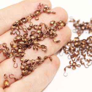 Peut inclure: Un grand tas de petits crochets en métal de couleur dorée avec une boule ronde à l'extrémité. Les crochets sont éparpillés sur une surface blanche.
