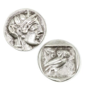 Może przedstawiać: Dwie srebrne monety z wypukłymi obrazami. Jedna moneta przedstawia profil kobiety w koronie. Druga moneta przedstawia sowę z rozpostartymi skrzydłami.