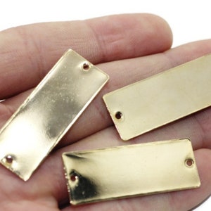 Può includere: Tre etichette rettangolari in metallo dorato con angoli arrotondati e due fori ciascuna. Le etichette sono appoggiate su una mano.