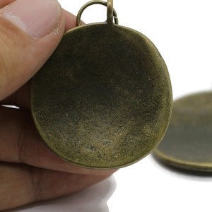 以下が含まれることがあります： 丸い形をしたアンティークゴールドの金属製のペンダントチャーム2個。チャームの上部にループが付いています。チャームは空白で、カスタマイズする準備ができています。