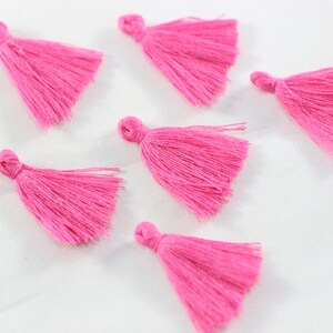 May include: Six pink fabric tassels with a knotted top and a fringe of fibres. The tassels are arranged in a scattered pattern on a white background.