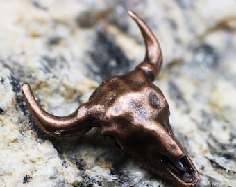 May include: A small, copper-colored bull skull sculpture with a detailed, textured surface. The sculpture is set against a backdrop of rough, light-colored stone.