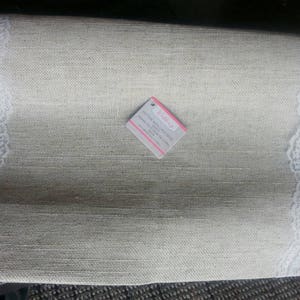 May include: A rectangular, beige linen placemat with white lace trim along two edges. A small tag with the word "wedding" is attached to the placemat. The placemat has a textured appearance.