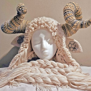 May include: A cream-colored crocheted ram's head hat with large, curved horns in shades of tan, gray, and blue. The hat features a braided fringe and decorative ear details. The hat is displayed on a white mannequin head.