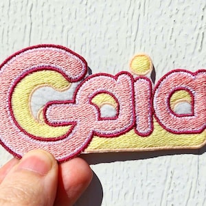 May include: A pink and yellow embroidered patch with the word "Gaia" in a retro font.