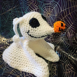 May include: A handmade crochet plush toy of Zero from The Nightmare Before Christmas. The white ghost dog has black eyes and a stitched smile. His nose is an orange pumpkin. The toy is set against a purple and silver spiderweb background.