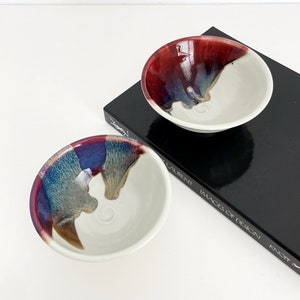 May include: Two small ceramic bowls with white interiors and colorful glaze exteriors. The bowls have a red, blue, and brown glaze on the outside. The bowls are sitting on a black book with white text that reads "Images of Design Knopf".
