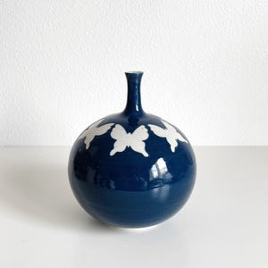 May include: A dark blue ceramic vase with a round body and a narrow neck. The vase is decorated with white butterfly silhouettes.