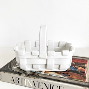 May include: White ceramic basket with a woven design and a handle. The basket is sitting on top of a book titled "Art in Venice".