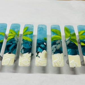 May include: Set of seven rectangular cabinet pulls. Each pull features a design with shades of blue, white, and green, resembling an abstract ocean scene with a touch of foliage. The pulls have a glossy finish.