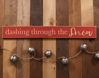 May include: A rectangular red wooden sign with the white text "dashing through the snow" engraved on the front. Below the sign, a piece of jute twine is strung with five metallic silver jingle bells, each featuring a star-shaped cutout.
